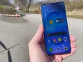 En hand håller en samsung liten mobil med sociala medier-appar. Utomhus med träd och en stig i bakgrunden.