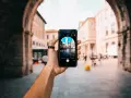 En hand håller upp en iPhone för att identifiera person på bild. Telefonen visar en livlig stadsgata med byggnader och människor.