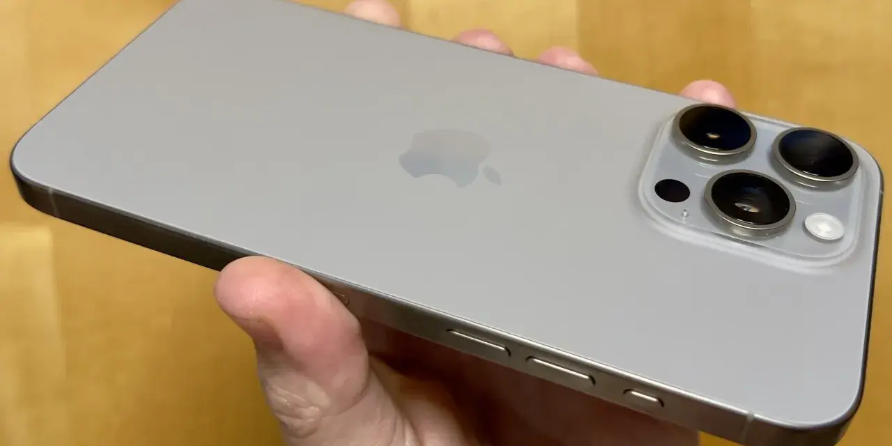 En hand håller en ljusgrå iPhone 15. Kameramodulen på baksidan har tre linser, redo att fånga dina bästa ögonblick.