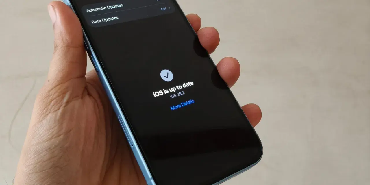 En hand håller en ljusblå iPhone. Skärmen visar "Software Update" och meddelandet "iOS is up to date" med version 26.2. En bekräftelse på att din ios uppdatering är klar.