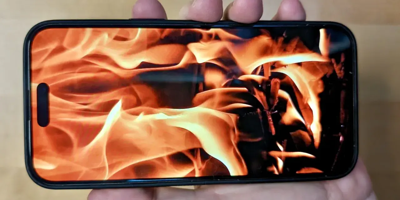 En hand håller en iPhone med en flammande eld som bakgrundsbild. Det ser ut som att iphone blir varm.