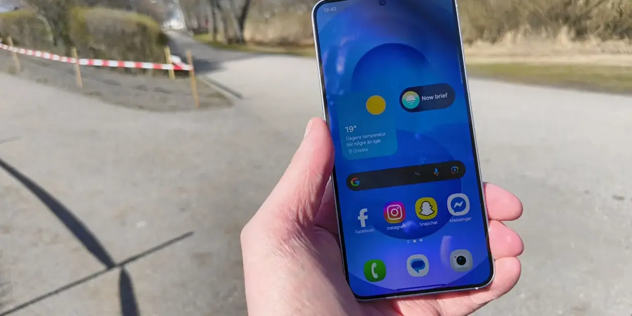 En hand håller en samsung liten mobil med sociala medier-appar. Utomhus med träd och en stig i bakgrunden.