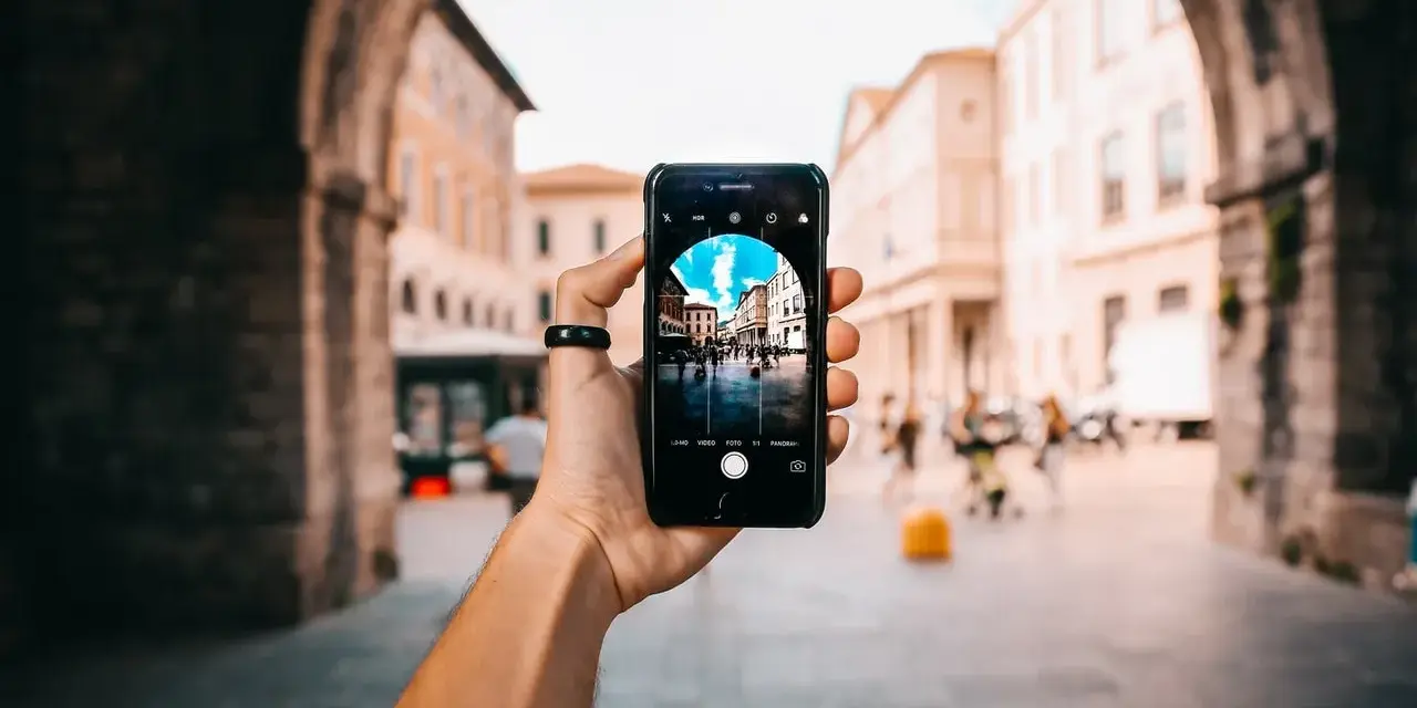 En hand håller upp en iPhone för att identifiera person på bild. Telefonen visar en livlig stadsgata med byggnader och människor.
