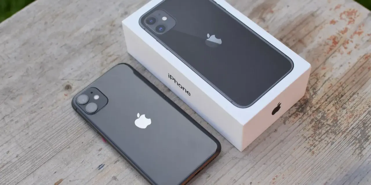 En mörkgrå iPhone 11 ligger bredvid sin vita förpackning på ett träbord. Perfekt iphone 11 storlek för handen.
