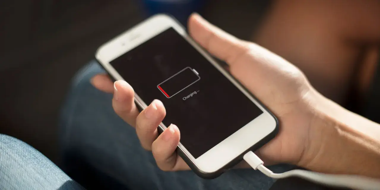 En hand håller en vit smartphone som laddas. Batteriikonen visar låg laddning och texten "Charging..." syns.