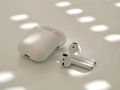 Vita trådlösa hörlurar och laddningsfodral på en ljus yta med solljus som skapar mönster. Perfekt för adaptivt ljud.