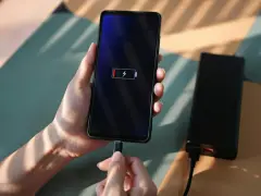 En hand håller en mobil som laddas med en datorladdare. Batteriikonen visar låg laddning.