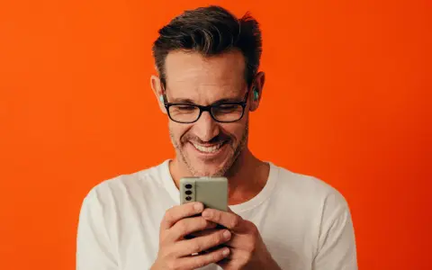 Man med glasögon och hörlurar ler när han tittar på sin iPhone.