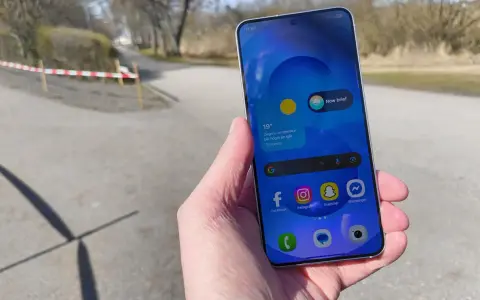 En hand håller en samsung liten mobil med sociala medier-appar. Utomhus med träd och en stig i bakgrunden.