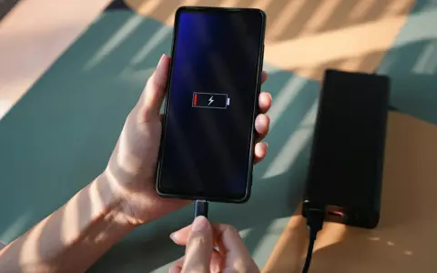 En hand håller en mobil som laddas med en datorladdare. Batteriikonen visar låg laddning.