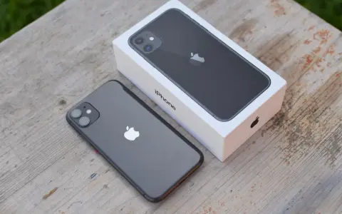 En mörkgrå iPhone 11 ligger bredvid sin vita förpackning på ett träbord. Perfekt iphone 11 storlek för handen.