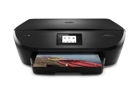 Svart HP Envy 5540-skrivare som skriver ut ett foto av färgglada båtar på en brygga. Enkel att skriva ut från iPhone.