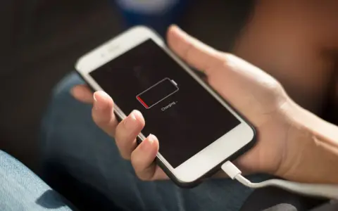 En hand håller en vit smartphone som laddas. Batteriikonen visar låg laddning och texten "Charging..." syns.