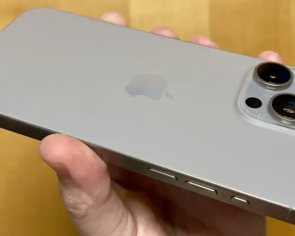 En hand håller en ljusgrå iPhone 15. Kameramodulen på baksidan har tre linser, redo att fånga dina bästa ögonblick.