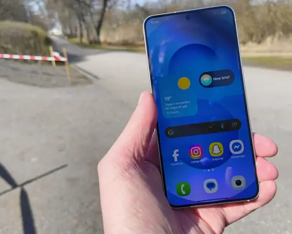 En hand håller en samsung liten mobil med sociala medier-appar. Utomhus med träd och en stig i bakgrunden.