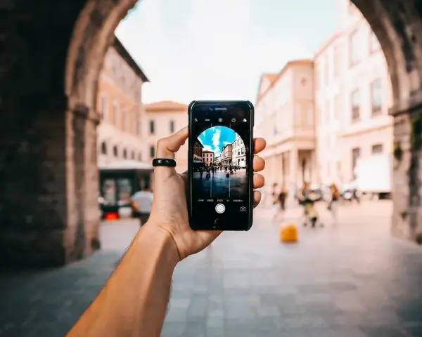 En hand håller upp en iPhone för att identifiera person på bild. Telefonen visar en livlig stadsgata med byggnader och människor.