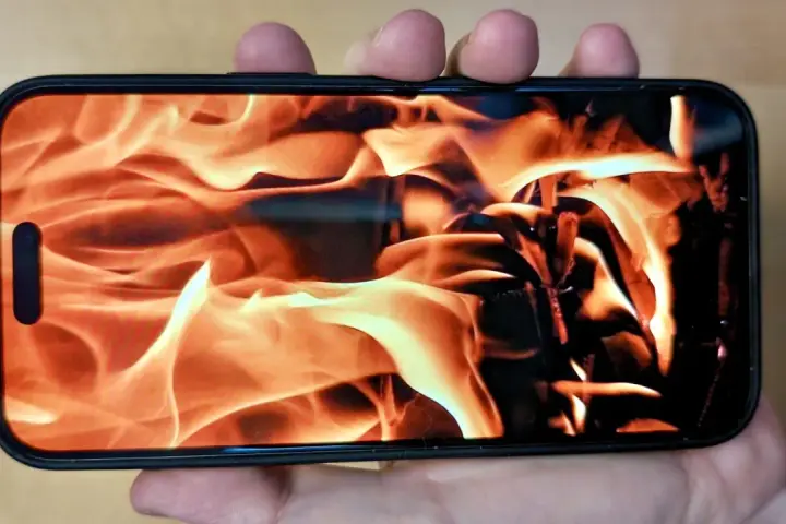 En hand håller en iPhone med en flammande eld som bakgrundsbild. Det ser ut som att iphone blir varm.