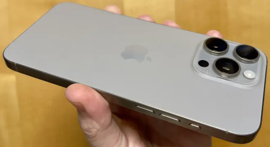 En hand håller en ljusgrå iPhone 15. Kameramodulen på baksidan har tre linser, redo att fånga dina bästa ögonblick.