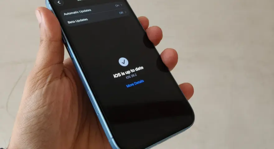 En hand håller en ljusblå iPhone. Skärmen visar "Software Update" och meddelandet "iOS is up to date" med version 26.2. En bekräftelse på att din ios uppdatering är klar.