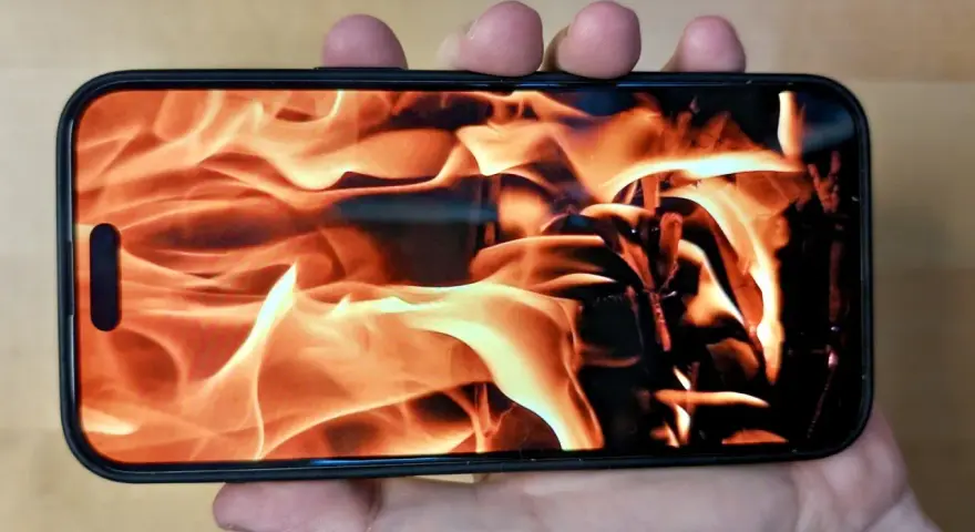 En hand håller en iPhone med en flammande eld som bakgrundsbild. Det ser ut som att iphone blir varm.