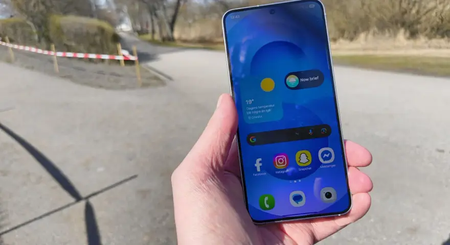 En hand håller en samsung liten mobil med sociala medier-appar. Utomhus med träd och en stig i bakgrunden.