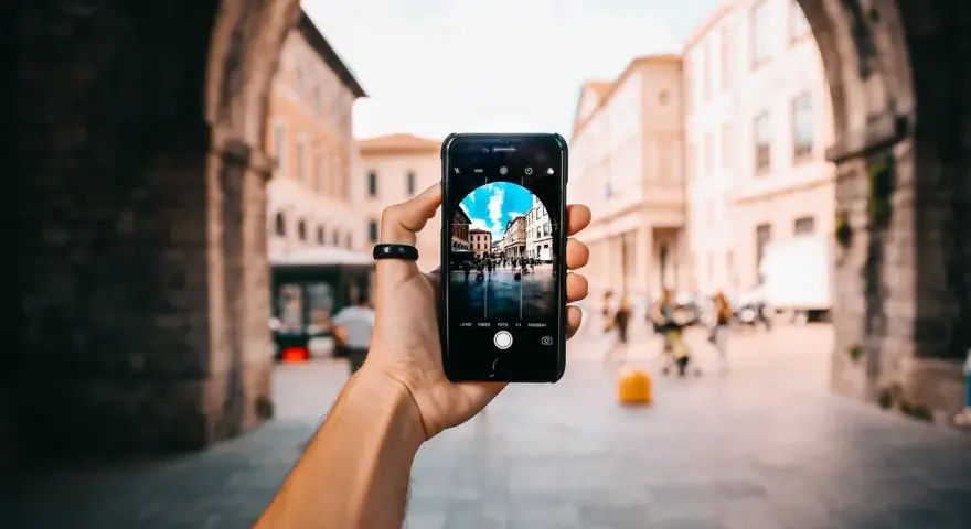 En hand håller upp en iPhone för att identifiera person på bild. Telefonen visar en livlig stadsgata med byggnader och människor.