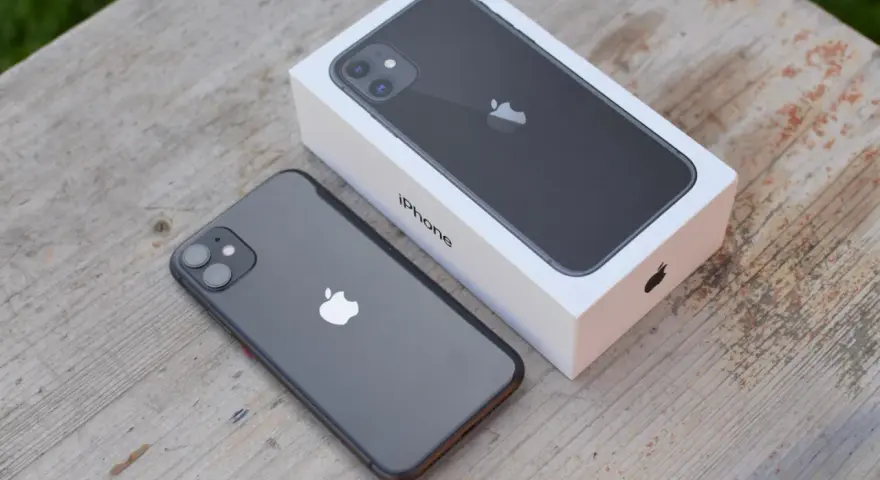 En mörkgrå iPhone 11 ligger bredvid sin vita förpackning på ett träbord. Perfekt iphone 11 storlek för handen.