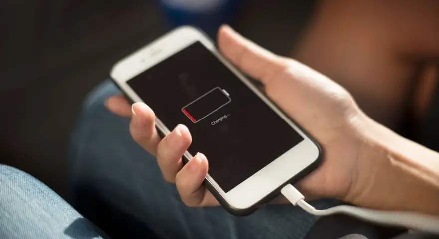En hand håller en vit smartphone som laddas. Batteriikonen visar låg laddning och texten "Charging..." syns.