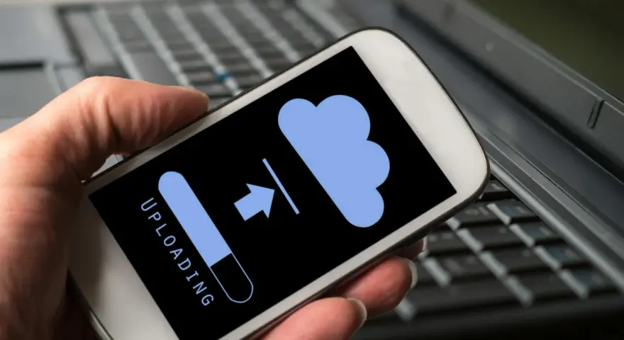 En hand håller en iPhone där texten "UPLOADING" visas med en laddningsindikator och en molnikon. Perfekt för att spara meddelanden iPhone i molnet.