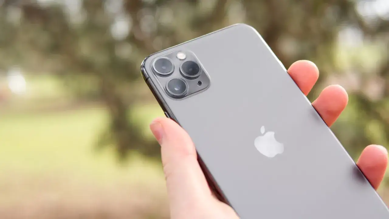 En hand h&aring;ller en gr&aring; iPhone 11. Kameramodulen med tre linser syns tydligt. Storleken p&aring; iPhone 11 &auml;r perfekt f&ouml;r att fotografera utomhus.