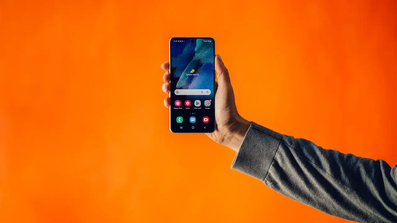 En hand håller upp en smartphone med dubbla simkort mot en orange bakgrund. Telefonen visar hemskärmen med appar.