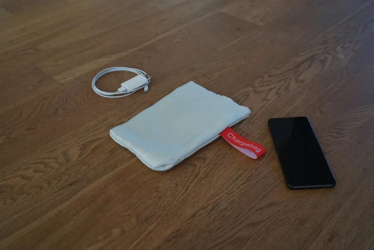 En mobil, laddare och en Chargebag p&aring; tr&auml;golvet. Perfekt f&ouml;r att ladda mobilen p&aring; natten utan att oroa sig f&ouml;r str&aring;lning.