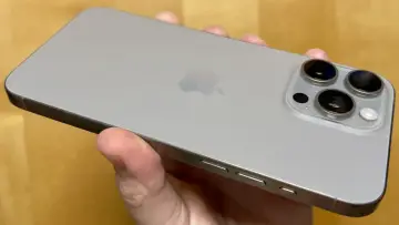 En hand håller en silverfärgad iPhone 15 Pro Max. Kameramodulen med tre linser syns tydligt.