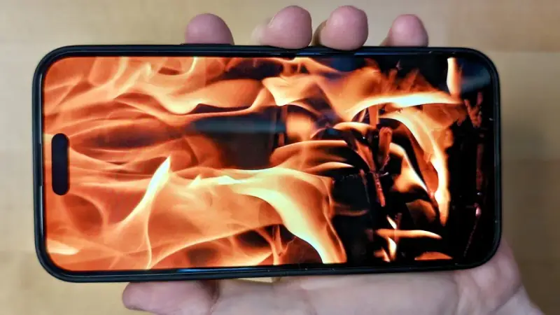 En hand håller en iPhone med en flammande eld som bakgrundsbild. Det ser ut som att iphone blir varm.